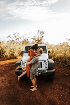 a man and woman kissing in front of a white jeep on dirt road next to tall grass