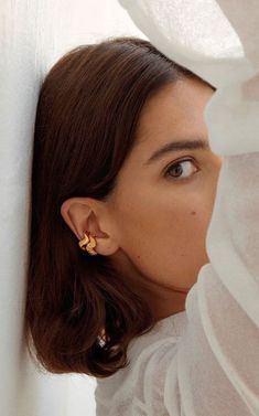 a woman wearing gold earring and white shirt looking out from behind the curtain with her eyes closed