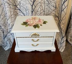 a white chest with flowers painted on the front and sides sitting on a wooden stand