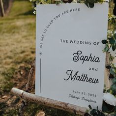 a wedding sign with flowers on it sitting in the grass next to a wooden fence