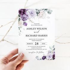 a person holding up a wedding card in front of a white background with purple flowers