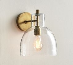 a clear glass light fixture mounted on a white wall with a gold metal hook and bulb