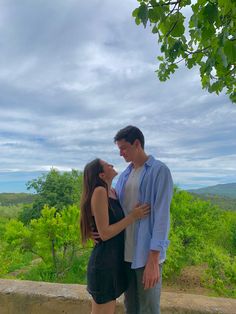 a man and woman standing next to each other on top of a hill with trees in the background