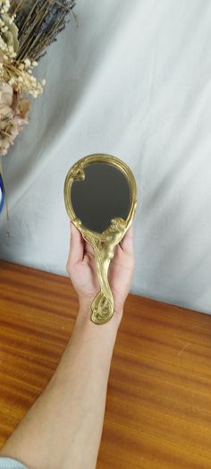 a person holding up a mirror on top of a wooden table next to a vase with dried flowers