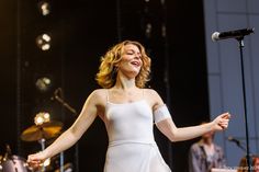 a woman in white dress standing on stage with her arms out and singing into a microphone