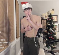 a shirtless man wearing a santa hat is standing in front of a christmas tree