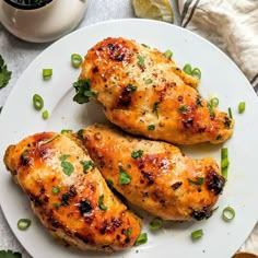 two pieces of chicken on a white plate with green onions and lemon wedges next to it