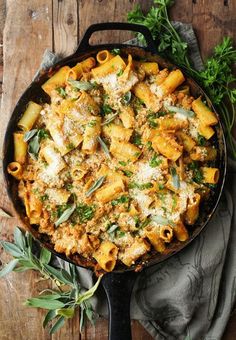 a skillet filled with pasta and sauce on top of a wooden table next to fresh herbs