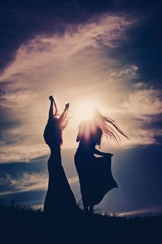 two women are dancing in the sun with their arms up and legs spread out, against a cloudy sky