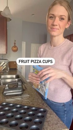 a woman standing in a kitchen holding a bag of fruit pizza cups next to a muffin pan