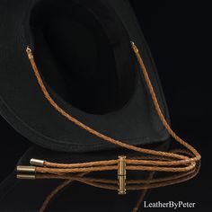 Enhance your cowboy hat with our premium whiskey-brown hand-braided stampede string or chin strap, meticulously crafted to complement Western-style hats. Crafted with versatility in mind, our strap can be effortlessly attached or fitted to your hat, ensuring a secure and stylish fit. Featuring an adjustable gold-tone metal slider, you can easily customize the size to your preference for optimal comfort. At each end of the braid, you'll find a golden metal aglet, adding a touch of refinement to t Luxury Leather Fedora For Western-themed Events, Rugged Leather Hats For Western-themed Events, Adjustable Leather Felt Hat For Western-themed Events, Brown Felt Hat For Western-themed Events, Artisan Brown Fedora For Western-themed Events, Brown Leather Hat, Cowboy Hat Bands, Custom Cowboy Hats, Leather Hat