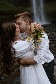 a man and woman are kissing in front of a waterfall while holding each other's shoulders