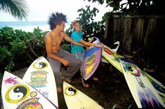 two people holding surfboards in front of some bushes and trees with the ocean in the background