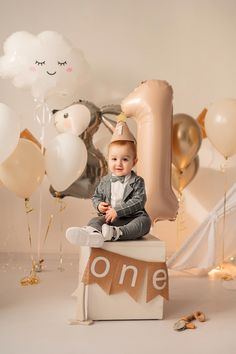 a little boy sitting on top of a cake with balloons around him and his first birthday sign