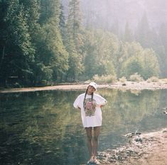 a woman standing on the shore of a lake wearing a white shirt and hat with trees in the background