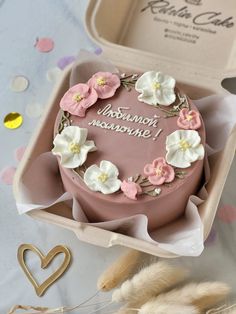 a pink cake with white and pink flowers on it in a box next to some feathers