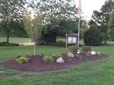 a flag pole in the middle of a flower bed with rocks and trees around it