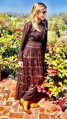 a woman standing on top of a brick walkway next to bushes and flowers in the background