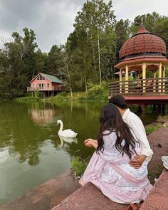 a man and woman sitting on the edge of a lake next to a gazebo