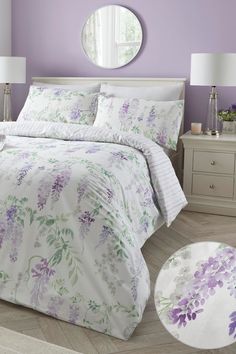 a white bed with purple flowers on it next to a round mirror and two lamps