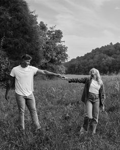 a man and woman holding hands while walking through tall grass in a field with trees behind them