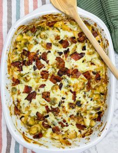 a casserole dish with bacon, cheese and broccoli in it next to a wooden spoon