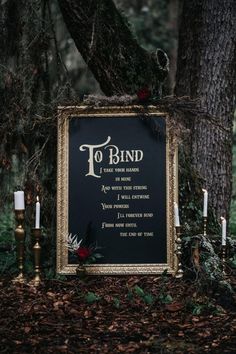 a black and gold framed sign sitting in the middle of a forest next to candles