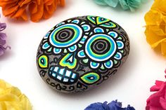 a painted rock surrounded by colorful flowers on a white surface with the colors of the day of the dead