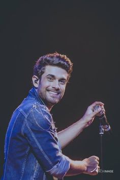 a smiling man holding a microphone in his right hand and wearing a denim jacket on