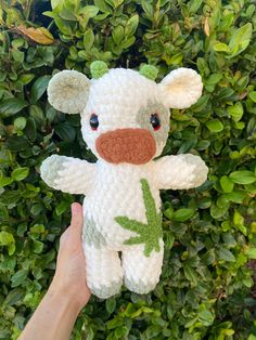 a hand holding a small white stuffed animal in front of some green leaves and bushes