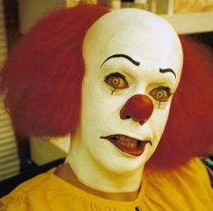 a creepy clown with red hair and white makeup looks at the camera while wearing a yellow shirt