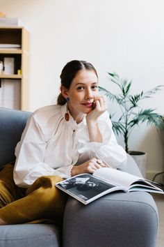 a woman sitting on a couch reading a book and looking at the camera with her hand under her chin