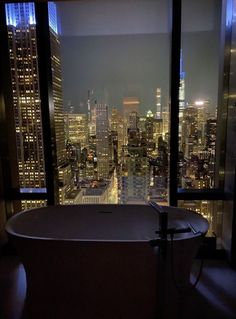 a bath tub sitting in front of a window overlooking a city at night with the lights on