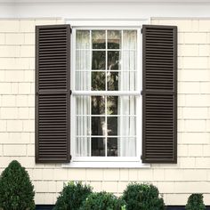 an open window with black shutters on the side of a white brick building next to shrubbery