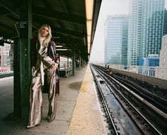 a woman leaning against a train station platform next to the rail road tracks with buildings in the background