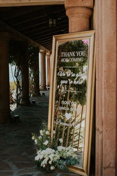 a large framed sign sitting on the side of a building next to flowers and greenery