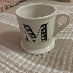 a white coffee cup with the letter m on it sitting on a quilted bedspread