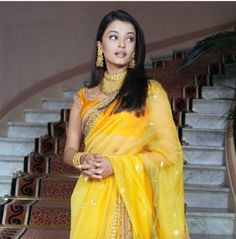 a woman in a yellow sari and gold jewelry standing on some stairs with her hand on her hip