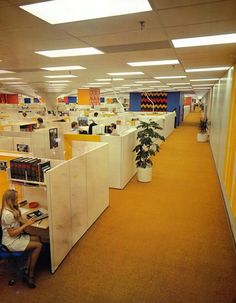 a woman sitting at a desk in an office cubicle with yellow walls and orange carpet