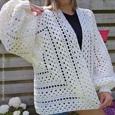 a woman wearing a white crocheted cardigan standing in front of a fence