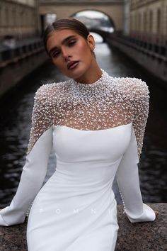 a woman in a white dress sitting on a ledge next to a river and looking at the camera