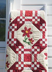 a red and white quilt hanging from the side of a house with flowers on it