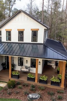 a white house with black metal roof and plants on the front porch, surrounded by trees