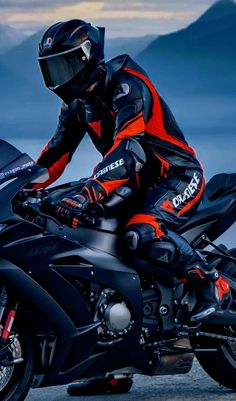 a man riding on the back of a black and orange motorcycle with mountains in the background