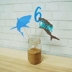 a mason jar filled with sand and two shark cutout sticks sitting on top of a wooden table