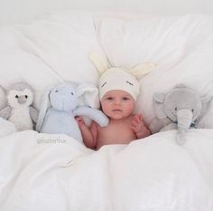 a baby laying in bed with stuffed animals on it's back and wearing a hat