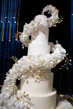 a three tiered white wedding cake with flowers on the top and sides, in front of a blue curtain