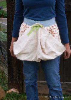 a woman standing in front of a fence wearing a white apron with green ribbon around the waist