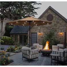 an outdoor fire pit with chairs and umbrella in front of a stone house at dusk