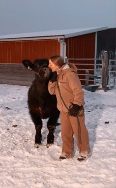 a woman standing next to a black horse on top of snow covered ground in front of a barn